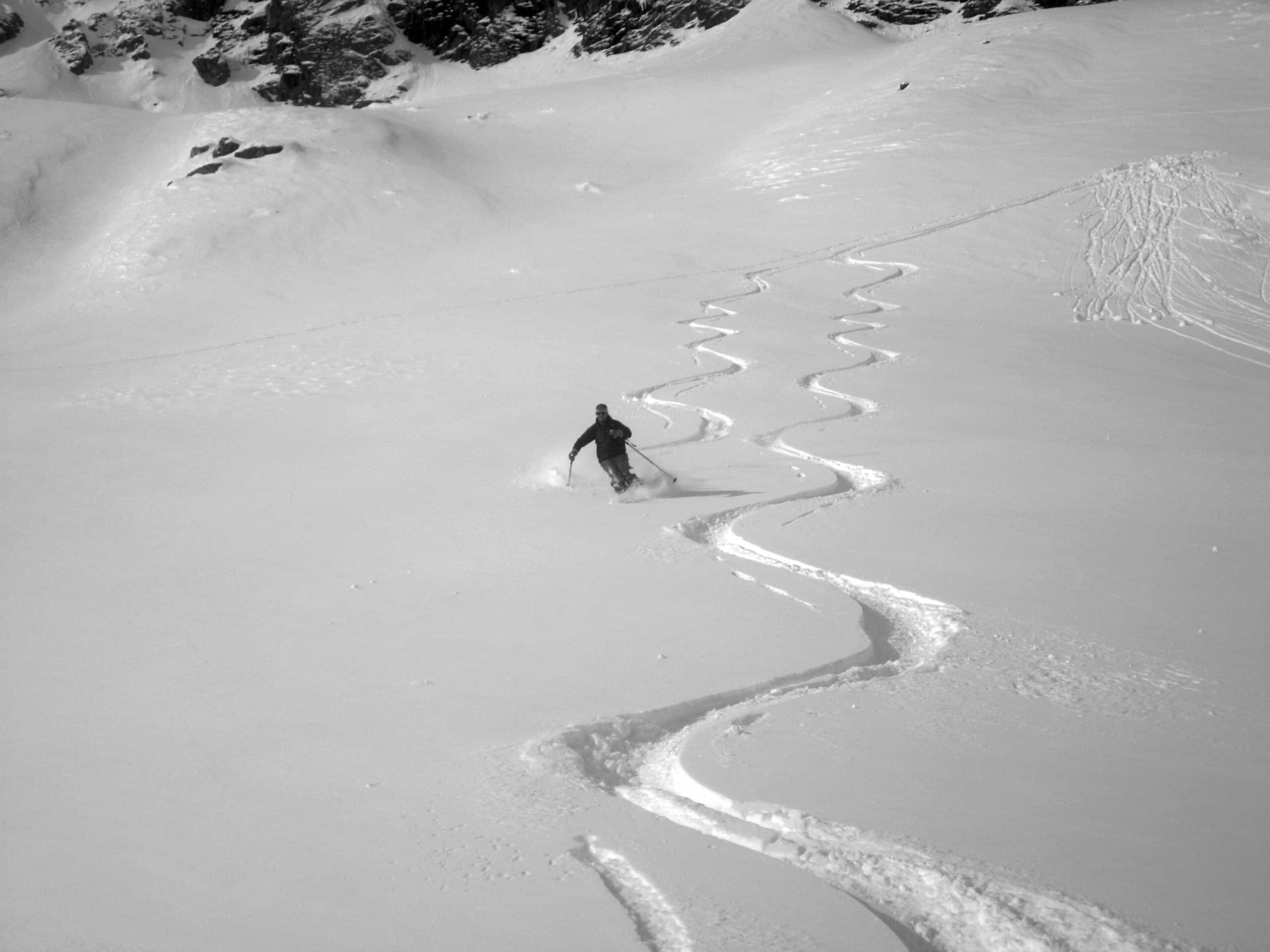discesa-scialpinismo-neve-fresca-powder Fotografia in bianco e nero di un momento di azione pura durante una discesa di scialpinismo in neve fresca powder. Uno sciatore traccia curve ampie e controllate su un pendio in pieno fuoripista, lontano dagli impianti di risalita.